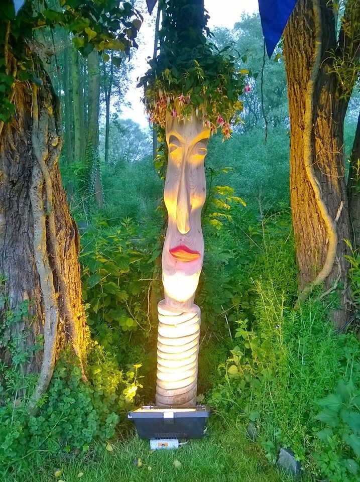 Visage de femme aux lèvres rouges sculpté dans un tronc, couronné de fleurs, éclairé le soir entre deux arbres.